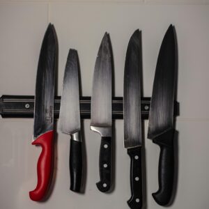 Five kitchen knives hanging on a magnetic strip on a white tiled wall.