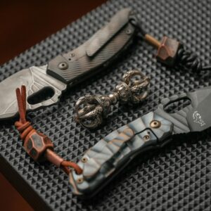 Stunning display of engraved pocket knives and ornate metal trinket on a textured black surface.